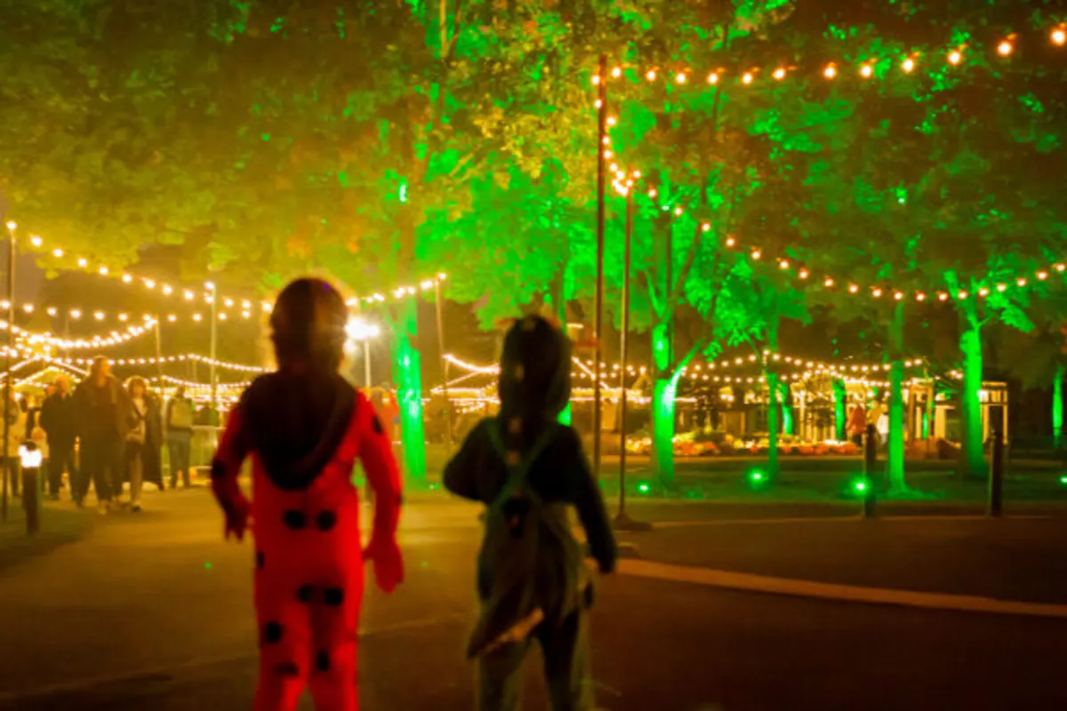 Two children in costumes walk under string lights and green-lit trees