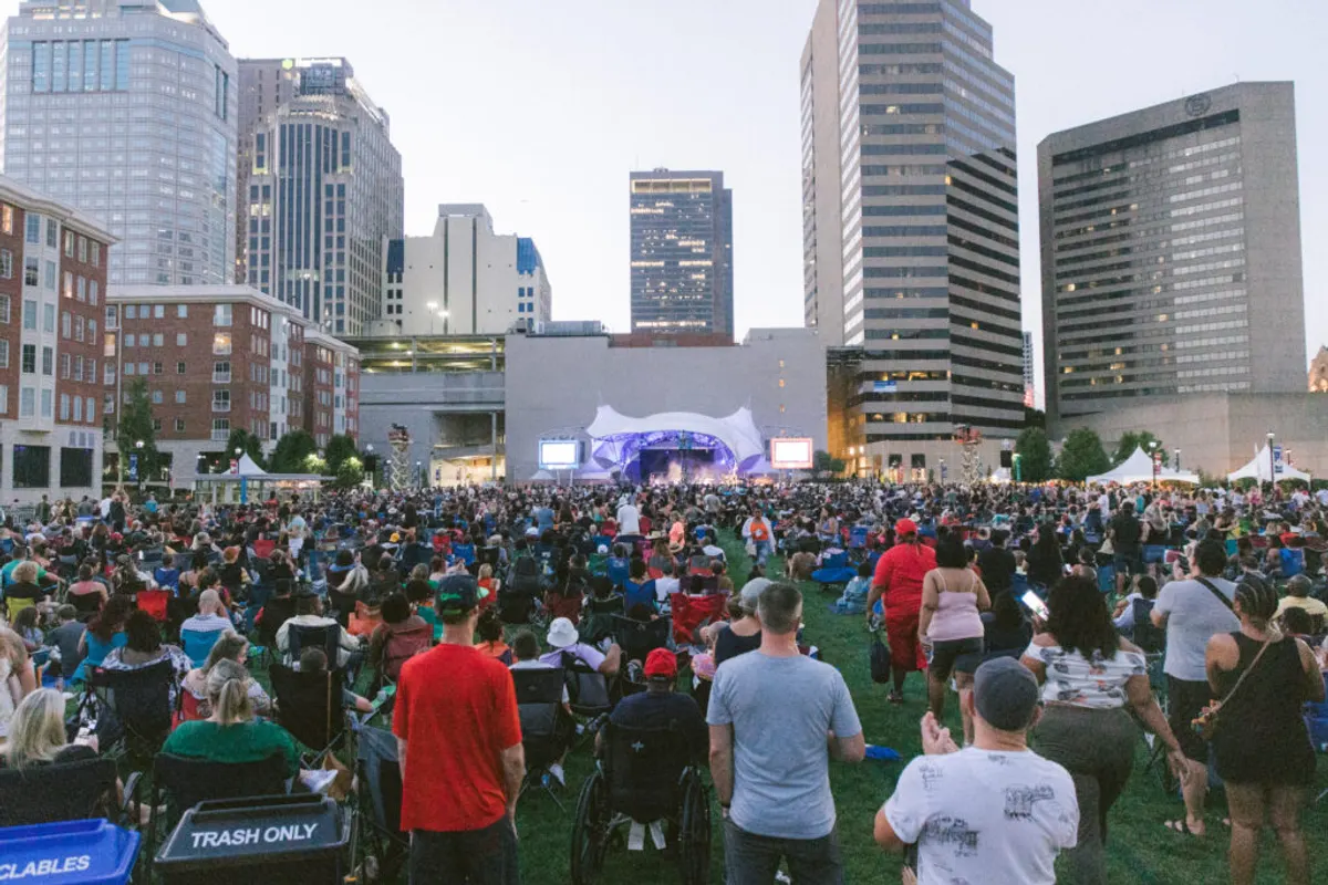 Large crowd on a lawn watching a stage beneath downtown towers