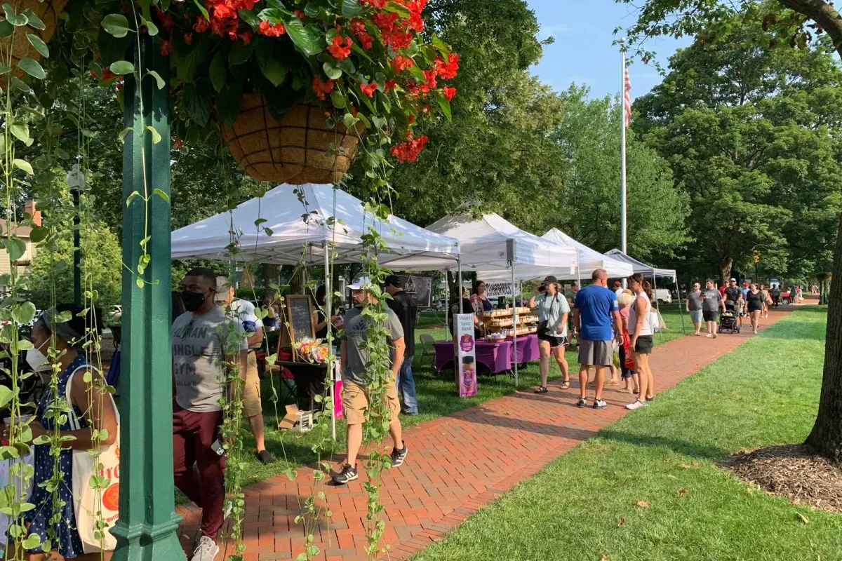 Shoppers walk a brick path past white vendor tents at a park market