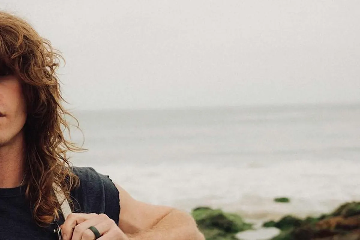 Jesse Welles with long hair and harmonica holder on a rocky beach