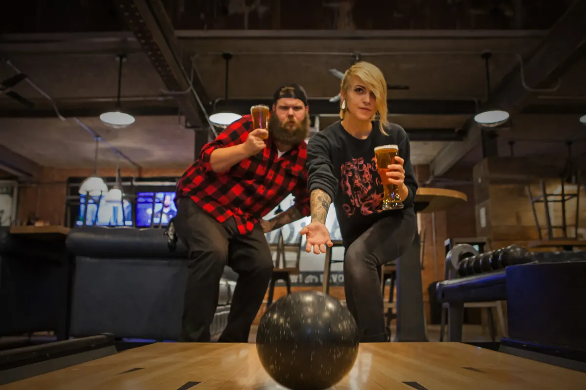 Two people holding beers about to roll a bowling ball